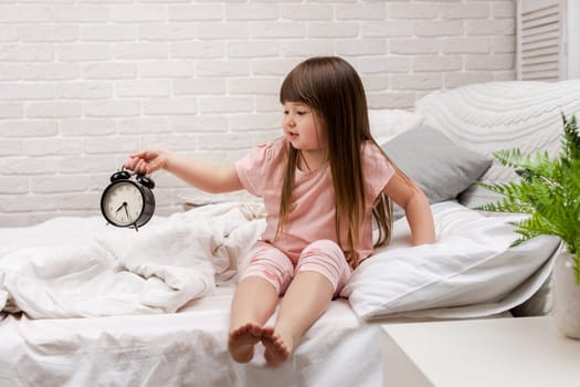 cute tired little child girl in pyjamas with clock in bed. good morning