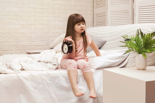 cute tired little child girl in pyjamas with clock in bed. good morning