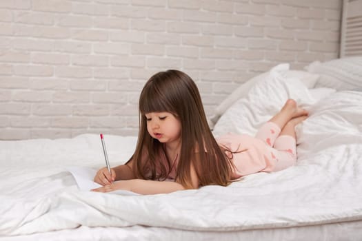 cute little girl drawing pictures while lying on bed. Kid painting at home