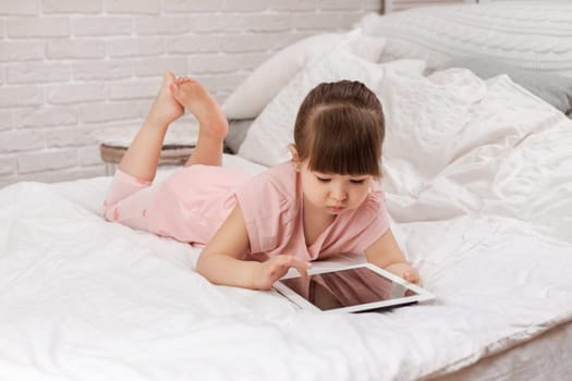 cute little child girl lies in bed uses digital tablet. child playing on tablet pc.