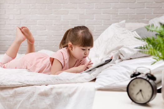 cute little child girl lies in bed uses digital tablet. child playing on tablet pc.