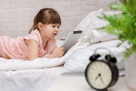 cute little child girl lies in bed uses digital tablet. child playing on tablet pc.