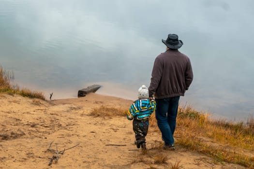 A happy family of two is standing on a cloudy autumn day on the bank of the lake and looking into the distance at the water surface, son and father stand with their backs to the camera