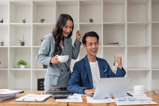 Two asian business people successful excited raised hands rejoicing with a laptop computer in office.