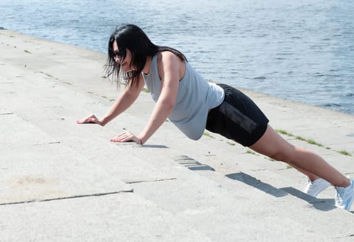 The girl is slim, tall and athletic in short shorts, doing push-ups on the street in the summer near the river. Strengthening the muscles of the arms and chest. Sports concept.