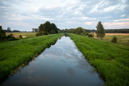 With a slow current, the river flows straight amidst green grasses and agricultural fields. The overcast sky is clearly reflected in the calm river.