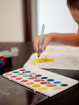 The boy paints with watercolors. Focus on case with many colors watercolors and blurred faceless baby boy draws on background.