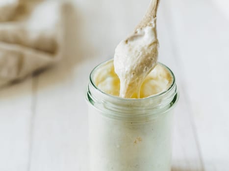 Ready for making bread wheat sourdough starter dripping from wooden spoon. Glass jar with sourdough starter 100 percent hydration on white wooden background. Copy space. Homemade breadmaking concept