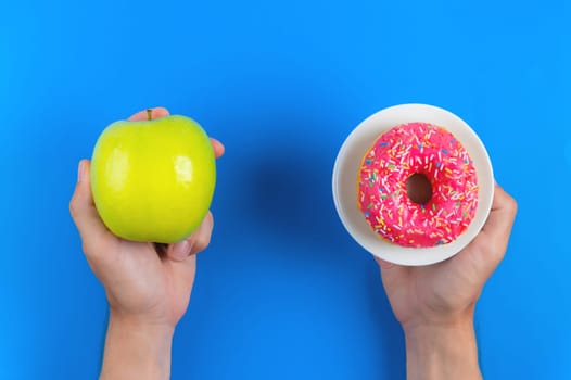 Green apple and pink donut in male hands, the concept of choosing health and unhealthy food on a blue background