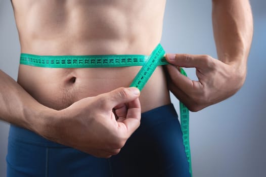 Close-up with waist and centimeter. The concept of a slim figure. Young fitness man measures his waist. Slim and athletic man in shorts checks his body. Body care. Weight loss and strong diet concept.