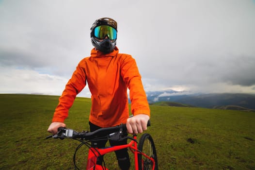 Portrait of a male downhill cyclist in a full face helmet and goggles with an orange windbreaker jacket with a raincoat against the backdrop of green hills and clouds over the mountains over the horizon. Looking at the camera