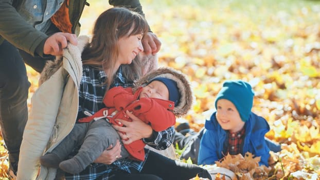 A mother and child in the park in the fall sitting on fallen leaves. Her husband and son come up behind her and hug her