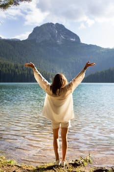 The girl stands with her back near a mountain lake in the background. Positive young woman traveling on a blue lake outdoors, adventure travel