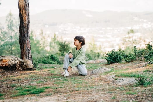 School boy kid relaxing sitting on the ground taking a breather during forest hiking, looking for dry sticks for the bonfire. Child resting on the ground in the city park