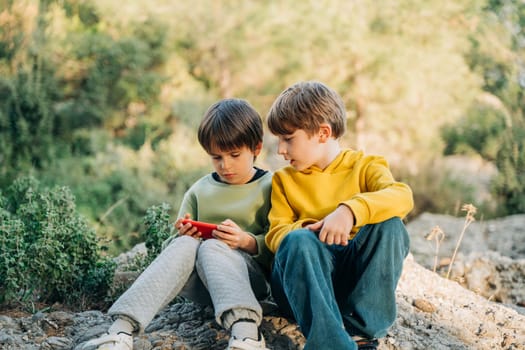 Elementary school kids friends playing a game on smartphone while sitting on a granite boulder in the city park. Children boys classmates watching gaming stream on a mobile cellphone in the forest.