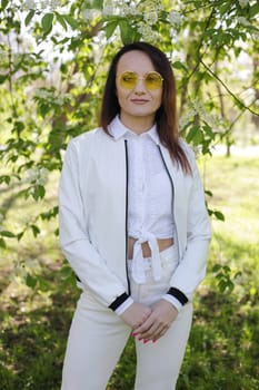 A beautiful woman in white clothes with yellow glasses and long hair. A positive woman with glasses on the background of a spring park.