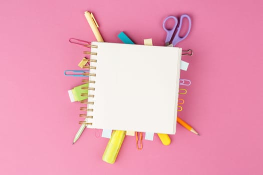 Open spiral notebook with bookmarks from paper clips and leaves for notes, pencil, scissors, binders on a pink background. Top view. Stationery concept. Office tools.