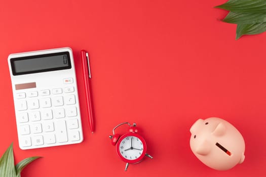 White calculator with a pen, alarm clock, with a pig piggy bank and green leaves on a red background. Top view.