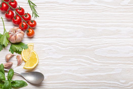 Healthy food ingredients composition with fresh cherry tomatoes, herbs, garlic cloves, lemon wedges and spoon on white wooden rustic background, overhead shot with copy space