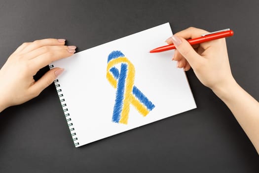 female hands draw yellow blue ribbon in notebook on dark table. concept needs help and support, truth will win