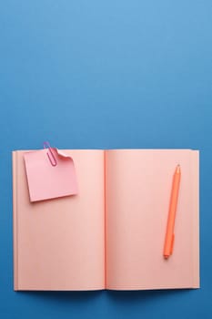 Pink notebook with pen, note sheet and color paper clip on blue isolated background. Flat lay. Top view. Vertical photo.