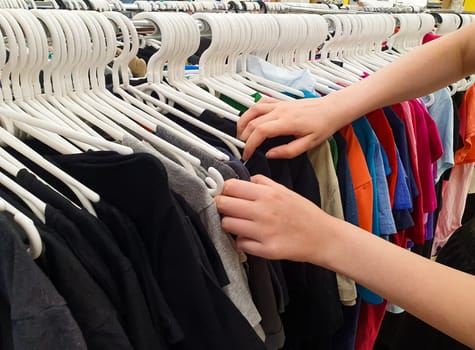 Women's hands are sorting through clothes on hangers. Second-hand. Old clothes are hung on hangers in the store.