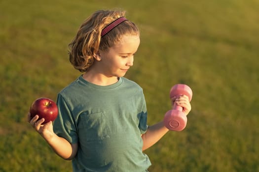 Healthy sporty child with apple and dumbbell outdoor in park. Cute child boy pumping up arm muscles with dumbbell. Fitness kids with dumbbells