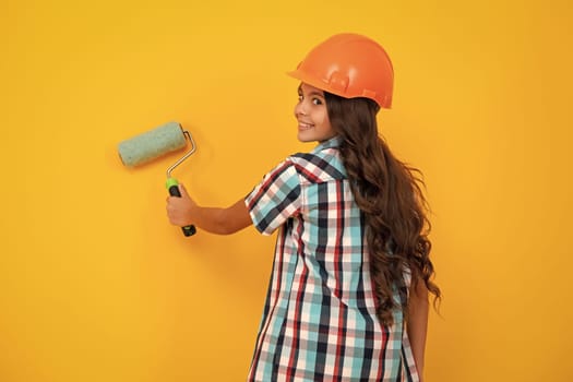 Child builder in hard hat helmet. Teenage girl painter with painting brush tool or paint roller. Teenager worker isolated on yellow background. Kids renovation concept