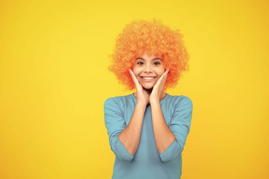 Birthday kids party. Funny kid in curly clown wig isolated on yellow background. Happy teenager portrait. Smiling girl