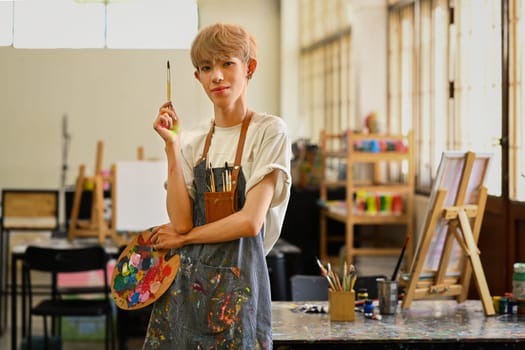 Portrait of asian gay man student wearing apron holding paintbrush and looking at camera.