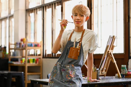 Young Asian gay man wearing apron holding paintbrush and smiling to camera. Art, education and leisure concept.