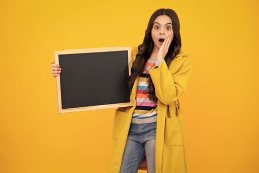 Teenage girl child holding blackboard, isolated on a yellow background. Shocked amazed face, surprised emotions of young teenager girl