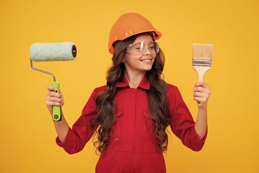 Child builder in helmet. Teenage girl painter with painting brush tool or paint roller. Child on repairing work isolated on yellow background. Kids renovation concept. Happy teenager girl