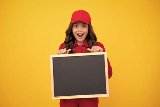 Excited child girl in red uniform and cap hold blackboard. Teenage girl hold blackboard isolated on yellow background. Copy space on empty board, mock up