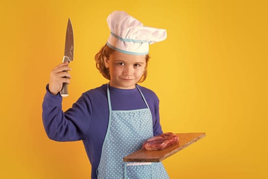Kid cook hold cutting board with meat beef steak and knife. Chef kid boy making healthy food. Portrait of little child in chef hat isolated on studio background. Child chef. Cooking process
