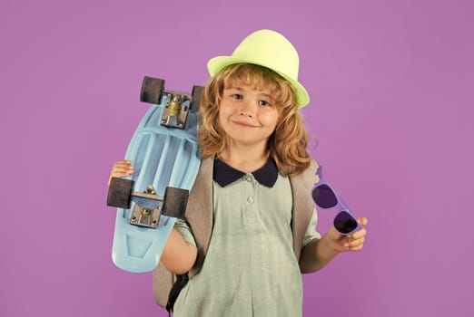 Child boy holding longboard on violet isolated background. Kid with pennyboard. Studio shot of cheerful little kid with penny board in fashion hat and summer shirt