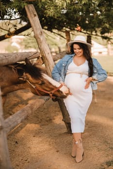 Stylish pregnant woman in a white hat in the countryside near a horse.