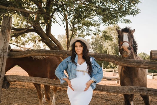 Stylish pregnant woman in a white hat in the countryside near a horse.
