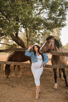 Stylish pregnant woman in a white hat in the countryside near a horse.