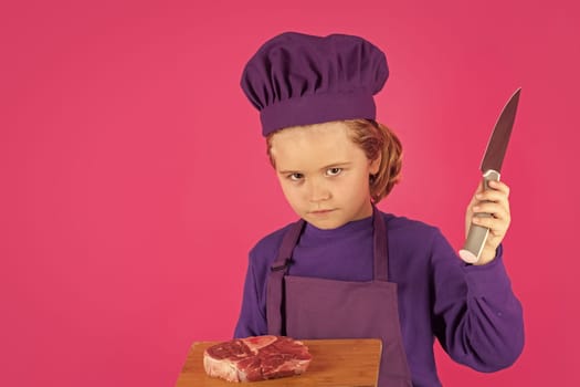 Child cook hold cutting board with meat beef steak and knife. Kid chef cook, studio portrait. Children cooking. Kid boy with apron and chef hat