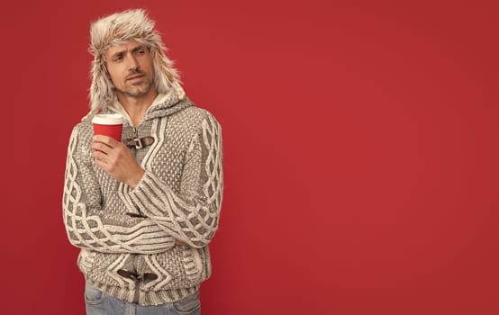 morning coffee in take away cup. serious grizzled guy in sweater and hat on red background. hoary man drinking coffee. male fashion model hold cup. winter fashion.