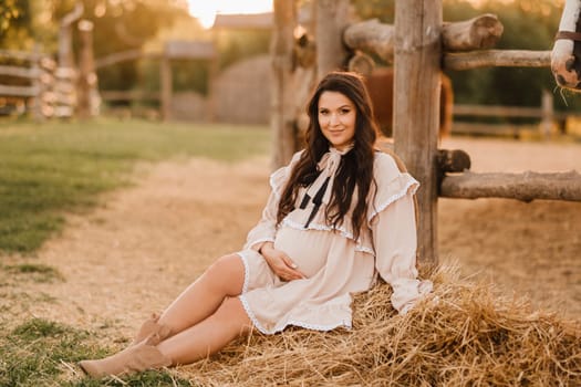 a pregnant woman in a dress in the countryside is sitting on the hay near the horses.