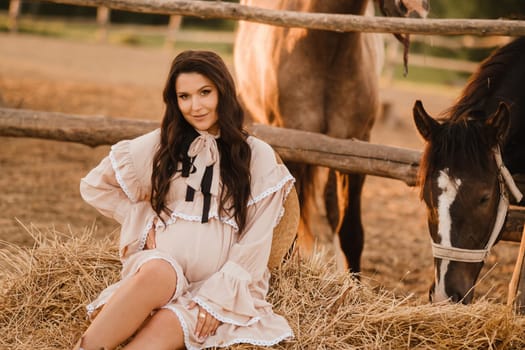 a pregnant woman in a dress in the countryside is sitting on the hay near the horses.