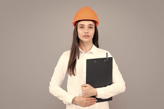 Serious woman worker in protective helmet with clipboard isolated on grey background. Young woman construction manager. Architect woman, female worker in hardhat helmet