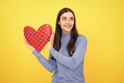 Romantic lovely girl. Young woman valentines day concept, girl with heart shaped card. Lovely charming girl holding in hands heart love symbol isolated on yellow background