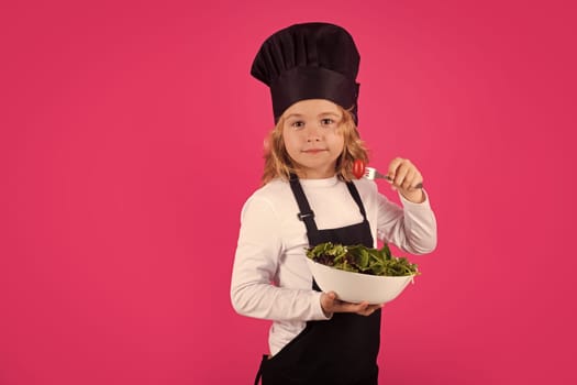 Kid cook hold plate with vegetables and fork with tomato. Cooking children. Chef kid boy making healthy food. Portrait of little child in chef hat isolated on studio background