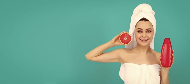 cheerful beautiful woman in towel with grapefruit shampoo bottle on blue background. Beautiful woman isolated face portrait, banner with mock up copy space