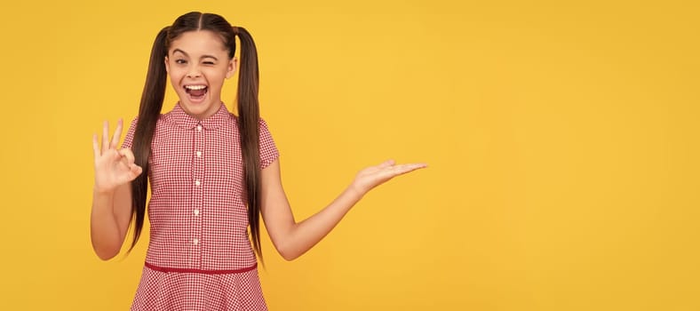 winking kid presenting product, copy space. ok. teen girl advertising on yellow background. Child face, horizontal poster, teenager girl portrait, banner with copy space