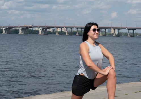 The girl is slender, tall and athletic in short shorts and sunglasses, in the summer she is engaged in fitness on the street by the river in the open air. Sports concept.