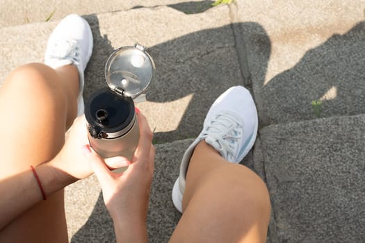 Sports concept. The girl sits on the steps in white sneakers and holds a bottle of water in her hand. Resting and quenches thirst after a workout on the street. No face. Close-up.